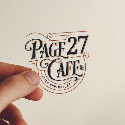 A hand holds a die-cut sticker featuring the text 'PAGE 27 CAFE' and 'ALICE SPRINGS, NT' in dark lettering with brown decorative outlines, against a plain off-white wall.